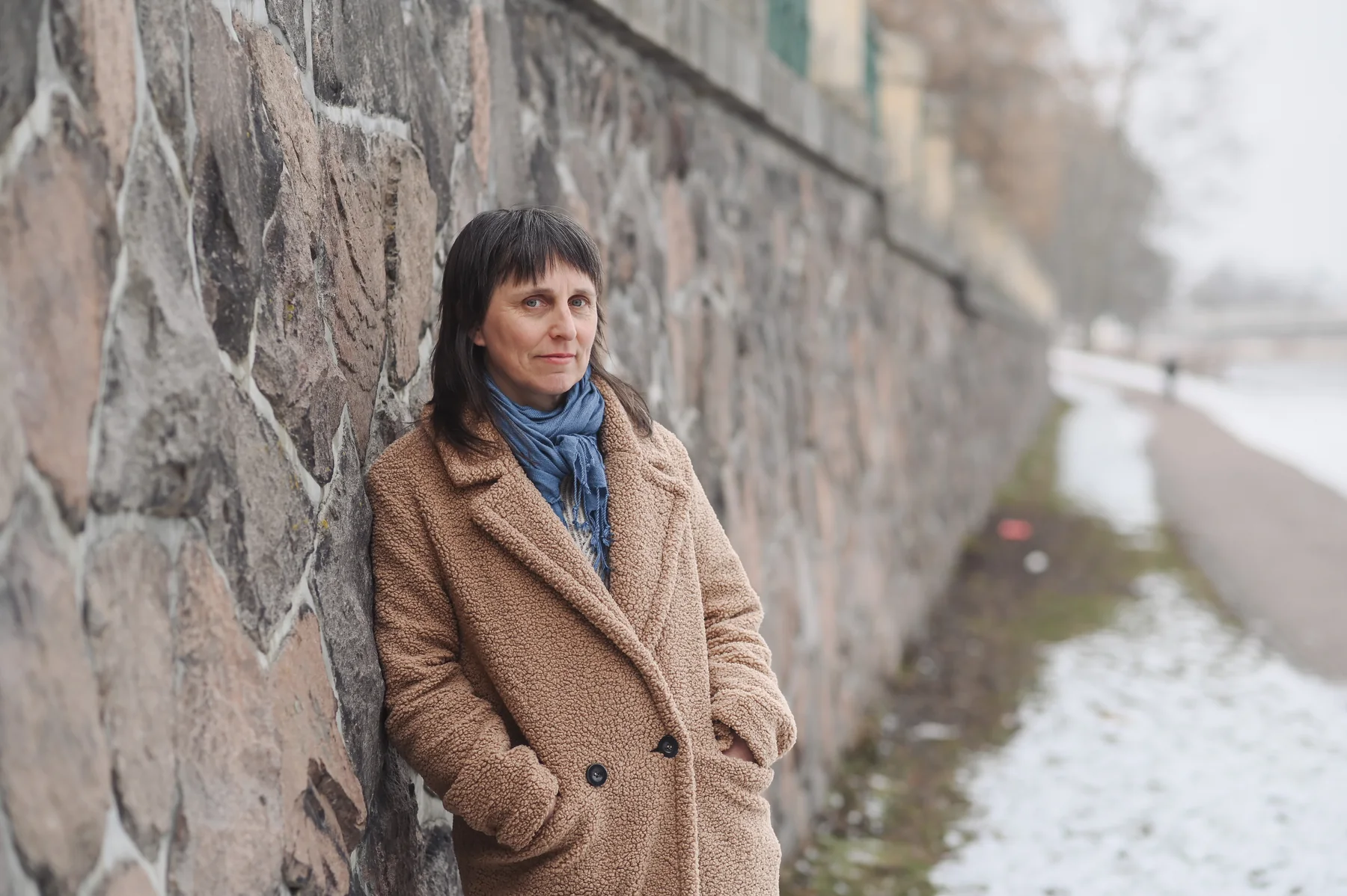Lucie Dlabolová v hnědém kabátu u kamenné zdi na náplavce